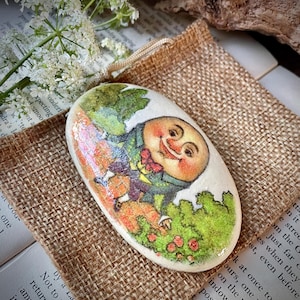 Op de afbeelding: Een ovale decoratieve steen met een Humpty Dumpty-illustratie. Het kunstwerk toont Humpty Dumpty in een groene jas en rode vlinderdas, tegen een achtergrond van groen gebladerte en oranje accenten. De steen ligt op een jute oppervlak.