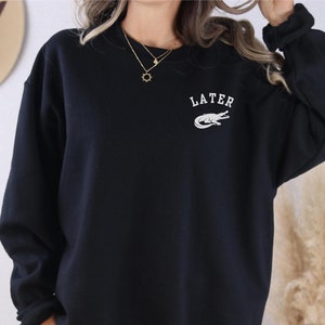 May include: Black sweatshirt with a white alligator graphic and the word "LATER" printed on the front.