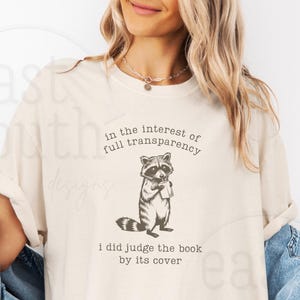May include: A light beige t-shirt featuring a raccoon graphic and the text "In the interest of full transparency, I did judge the book by its cover."