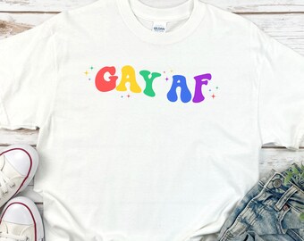 May include: White t-shirt with the text "GAY AF" in rainbow colors and glitter accents.