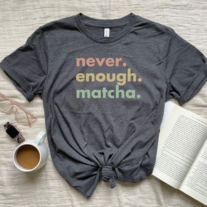 以下が含まれることがあります： 虹色のカラーで「never. enough. matcha.」という文字がプリントされたダークグレーのTシャツ。Tシャツは裾で結ばれています。