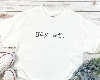 May include: White t-shirt with the text "gay af." printed in black on the front. The shirt is laid flat on a white wooden surface with a pair of white sneakers and blue jeans.