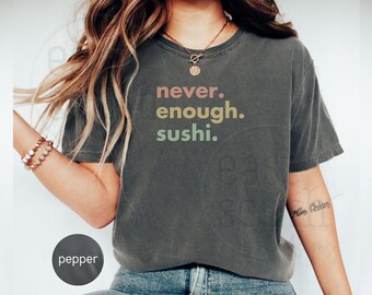 May include: A dark grey cropped t-shirt with the text "never. enough. sushi." printed in colourful letters. The shirt is being worn by a person with long brown hair.