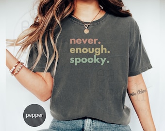 May include: Dark gray t-shirt with the words "never. enough. spooky." in a colorful, layered font. The shirt has a classic crew neck and short sleeves. The shirt is made of a soft, comfortable material.