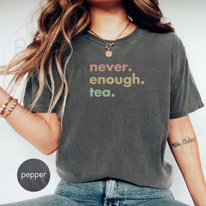 以下が含まれることがあります： 暗いグレーのTシャツに「never. enough. tea.」という文字がカラフルな文字でプリントされています。