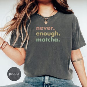 以下が含まれることがあります： レインボーカラーで「never. enough. matcha.」とプリントされたダークグレーのクロップドTシャツです。