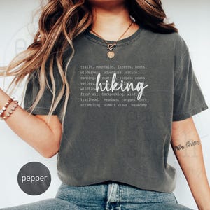 Könnte beinhalten: Ein dunkelgraues T-Shirt mit einem weißen Grafikdesign, das "trails, mountains, forests, boots, wilderness, adventure, nature, camping, valleys, ridges, peaks, wildflower, fresh air, backpacking, wilderness, trailhead, meadows, canyons, rocks, scrambling, summit views, basecamp" lautet, wobei das Wort "hiking" in einer größeren Schriftart dargestellt ist.