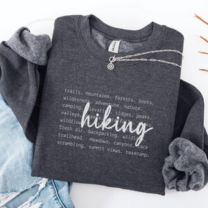 May include: A dark gray sweatshirt with a white graphic design that defines "hiking" as "trails, mountains, forests, boots, wilderness, adventure, nature, camping, elevation, ridges, peaks, valleys, trees, waterfalls, wildflowers, fresh air, backpacking, wildlife, trailhead, meadows, canyons, lock, scrambling, summit views, basecamp."