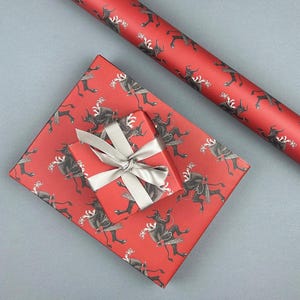 May include: Red wrapping paper and a wrapped gift with a silver bow. The paper and gift are decorated with a repeating pattern of black figures. A roll of matching wrapping paper is also visible.