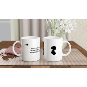 Puede incluir: Dos tazas de cerámica blanca. Una tiene el texto negro "I prefer to be unsociable and taciturn". La otra presenta una silueta negra de una mujer. Las tazas están sobre una superficie a cuadros.