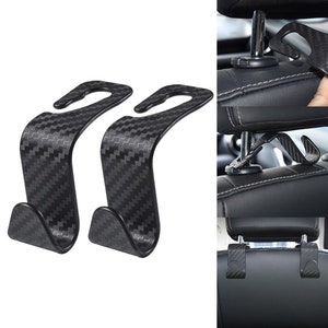May include: Two black car seat headrest hooks with a carbon fiber pattern. The hooks are designed to hang bags, coats, or other items on the back of a car seat.