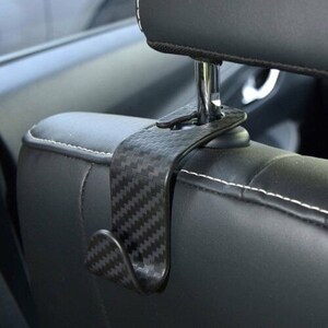 May include: Black car seat hook with a carbon fiber pattern. The hook is designed to hang on the back of a car seat.