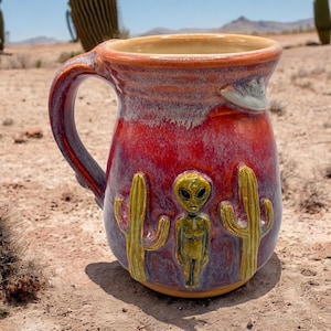 May include: A ceramic mug with a glossy red and purple glaze featuring a raised, sculpted green alien figure standing between two green saguaro cacti. A small, sculpted flying saucer is positioned near the rim of the mug, which has a handle.