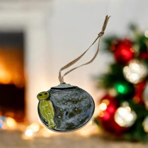 May include: A ceramic Christmas ornament with a gray speckled finish and a green alien figure. The ornament is round with a brown string for hanging. Festive holiday decor.