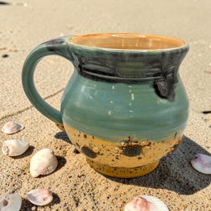 May include: A ceramic mug with a rounded body and a curved handle. The upper portion features a glossy teal glaze with dark drips, while the lower section is a speckled tan color with small, raised, dark brown circular textured accents.