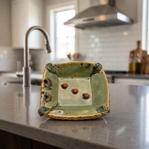 May include: A square ceramic bowl with a glossy sage green glaze, featuring three small red berry-like embellishments in the center and dark green leaf-shaped accents at each of the four corners, with a textured, uneven rim and base.