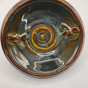 May include: A round ceramic bowl featuring a glossy glaze in shades of deep blue, green, and brown. The center displays a spiral pattern, and two raised, copper-colored dragonfly figures are positioned on opposite sides of the interior.