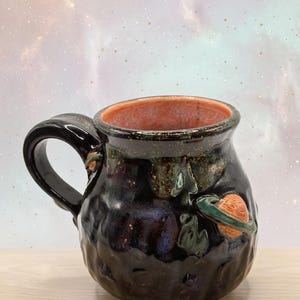 May include: A round ceramic mug with a glossy black glaze and a textured, dimpled surface. The interior is a warm terracotta orange. A small, sculpted orange planet with a green ring is attached to the side, featuring a dark, curved handle.