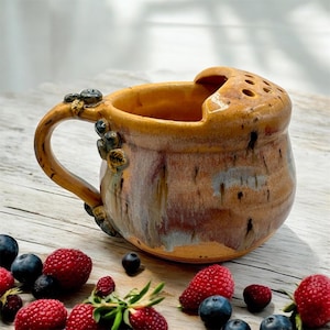 May include: A handmade ceramic mug with a unique design. The mug is primarily brown with orange and blue accents. It features a handle adorned with small, dark blue embellishments. The top has a built-in flower frog with multiple holes. Fresh raspberries and blueberries surround the mug.