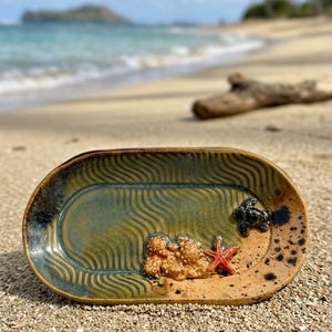 May include: An oval ceramic tray with a green and tan glaze featuring a wavy texture. The tray is decorated with a small, raised, dark-colored sea turtle, a cluster of textured beige coral, and a bright orange starfish on the surface.