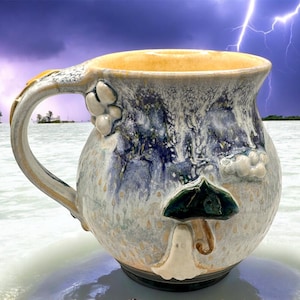 May include: Handmade ceramic mug with a mushroom design. The mug features a cream-colored base with blue and white accents, a yellow interior, and a decorative handle. A green and white mushroom is sculpted on the front. The mug is approximately 5 inches tall.