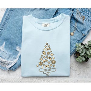 May include: A light blue t-shirt featuring a graphic of a Christmas tree composed of various gaming icons, including handheld consoles, game controllers, and polyhedral dice. The shirt has a crew neckline and a Comfort Colors brand label.