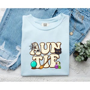 Pode incluir: Uma t-shirt azul-clara com a palavra AUNTIE em letras grandes bege com padrão de estrelas. O design inclui uma teia de aranha com uma aranha, um gato preto, uma bola de cristal, uma vassoura e dois pequenos frascos de poção roxo e verde.