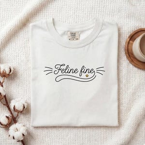 May include: A white t-shirt featuring the black text Feline fine in a cursive script, accented by three small lines on each side and a tiny yellow star. The shirt has a crew neckline and a Comfort Colors brand label visible at the inner collar.