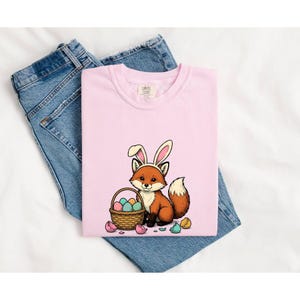 Puede incluir: Una camiseta rosa claro con un gráfico de un zorro marrón que lleva orejas de conejo blancas y rosas, sentado junto a una cesta de mimbre llena de huevos de Pascua de colores, con cáscaras de huevo rotas esparcidas por el suelo.