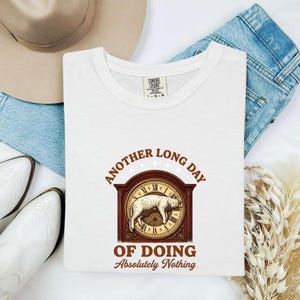 May include: A white t-shirt featuring a graphic of a cat sleeping on a clock face with the text "ANOTHER LONG DAY OF DOING Absolutely Nothing" in brown lettering. The shirt has a Comfort Colors brand label on the inside of the neckline.