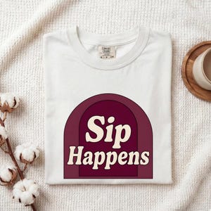 Puede incluir: Una camiseta blanca de manga corta con un gráfico de arco granate y el texto de color crema "Sip Happens" centrado en la parte delantera. La camiseta tiene un cuello redondo y una etiqueta de la marca Comfort Colors visible en el interior.