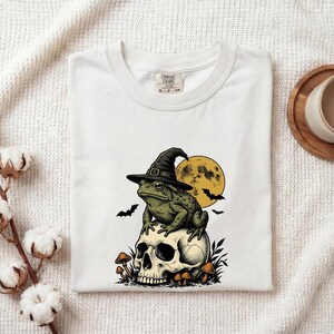 May include: A white t-shirt featuring a graphic of a green toad wearing a black witch hat, perched atop a human skull. The design includes a yellow full moon, silhouettes of flying bats, and small mushrooms at the base of the skull.