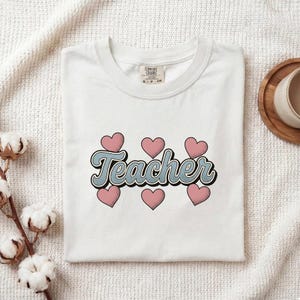 May include: A white short-sleeved t-shirt featuring the word Teacher in a blue script font with a black outline. The text is surrounded by six pink heart shapes arranged in two rows above and below the word. The shirt has a Comfort Colors label.