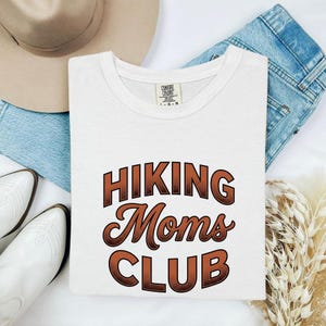 以下が含まれることがあります： 茶色のグラデーションフォントで「HIKING Moms CLUB」という文字がプリントされた白い半袖Tシャツ。内側の襟元にはComfort Colorsのブランドラベルが付いており、ベージュの帽子、青いデニムパンツ、白いブーツと一緒に配置されています。
