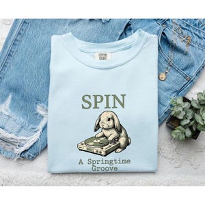 May include: A light blue crewneck t-shirt featuring a graphic of a rabbit using a turntable. The text above the rabbit reads SPIN, and the text below reads A Springtime Groove. The shirt has a Comfort Colors brand label at the inner neckline.