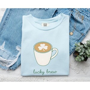 Puede incluir: Una camiseta de cuello redondo azul claro con un gráfico de una taza de café blanca con un diseño de trébol en la espuma. El texto "lucky brew" está impreso en verde debajo de la taza. La camiseta tiene una etiqueta Comfort Colors en el cuello.