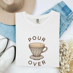 Op de afbeelding: Een wit t-shirt met een grafische print van een koffiefilterhouder en de tekst "POUR OVER" in bruine letters. Het shirt is voorzien van een Comfort Colors merklabel bij de binnenhalslijn. Het ontwerp is gecentreerd op de borst.