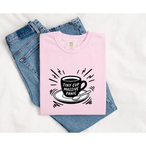 Może przedstawiać: Jasnoróżowy t-shirt z czarną grafiką przedstawiającą filiżankę kawy na spodku z łyżeczką. Napis na filiżance brzmi "TINY CUP MASSIVE PANIC". Koszulka leży na niebieskich dżinsach. Wzór zawiera promieniście rozchodzące się linie.
