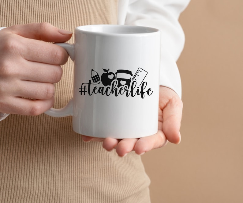 May include: White ceramic mug with a black design of a pencil, apple, and ruler with the text "#teacherlife" on it.