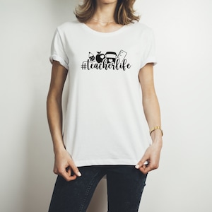 May include: White t-shirt with a black graphic of a pencil, apple, book, and ruler with the text "#teacherlife" printed on the front.