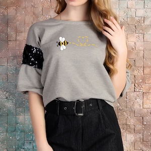 May include: Gray short-sleeved shirt with a bee graphic and black sequins on the sleeves. The bee is flying and leaving a trail of hearts behind it.