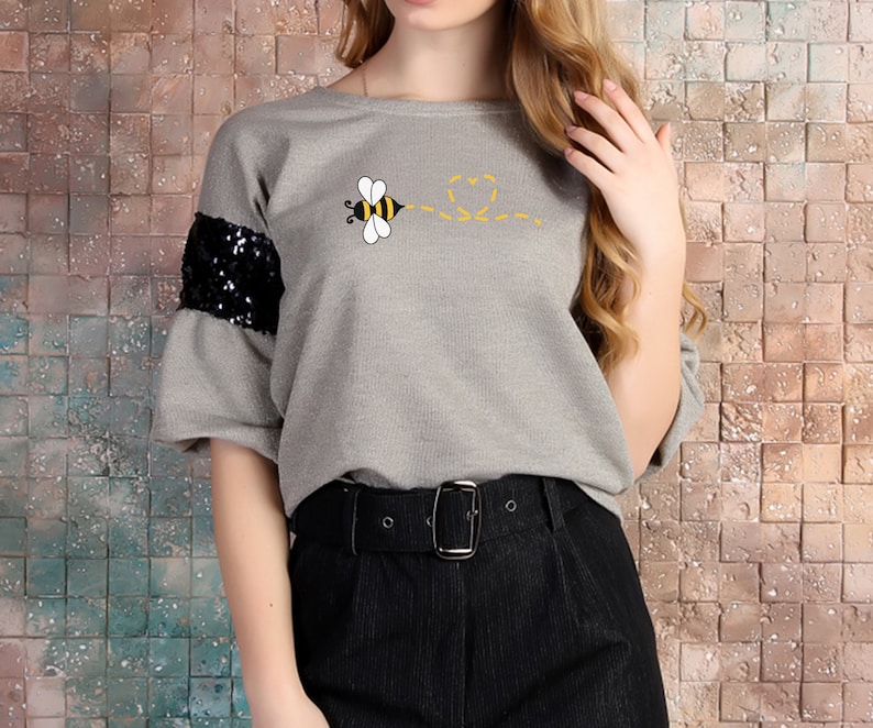 May include: Gray short-sleeved shirt with a bee graphic and black sequins on the sleeves. The bee is flying and leaving a trail of hearts behind it.