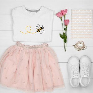 May include: A white t-shirt with a yellow and black bee graphic, a pink tulle skirt with gold stars, white sneakers, a pink and white striped notebook, and pink flowers.