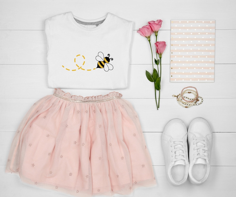 May include: A white t-shirt with a yellow and black bee graphic, a pink tulle skirt with gold stars, white sneakers, a pink and white striped notebook, and pink flowers.