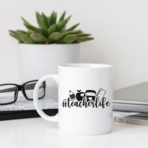 May include: White ceramic coffee mug with a black design of a pencil, apple, coffee cup, and ruler with the text "#teacherlife" printed on the side.