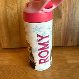 May include: A white water bottle with a pink lid and handle. The bottle features a design with snowflakes and a cartoon character. The name "ROMY" is printed vertically in pink.