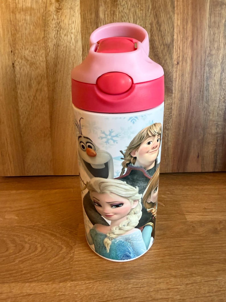 May include: A pink and red water bottle featuring characters from the animated movie Frozen. The bottle has a flip-top lid and a white body with images of Elsa, Anna, Olaf, and Kristoff. The bottle is designed for kids.