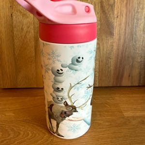 May include: A white water bottle with a pink lid and a red band. The bottle features a cartoon design with snowflakes, a snowman, and a reindeer. The bottle is designed for kids.