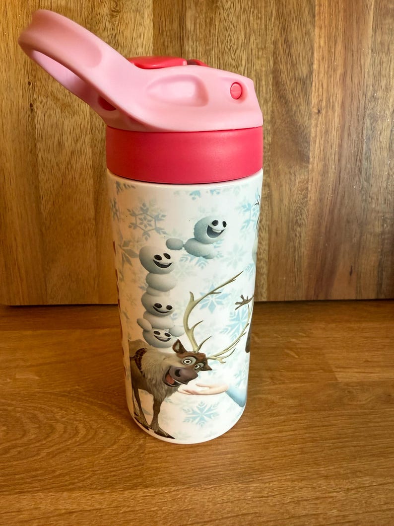 May include: A white water bottle with a pink lid and a red band. The bottle features a cartoon design with snowflakes, a snowman, and a reindeer. The bottle is designed for kids.