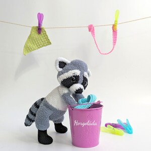 K&ouml;nnte beinhalten: Ein geh&auml;keltes Waschb&auml;r-Spielzeug, das ein blaues geh&auml;keltes Seil in einem rosafarbenen Eimer mit dem Wort "Horgol&aacute;da" auf der Seite h&auml;lt. Der Waschb&auml;r steht auf einer wei&szlig;en Oberfl&auml;che mit einem Garnfaden, der dar&uuml;ber h&auml;ngt.